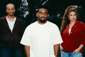 Three people standing outdoors, with the center person in a white shirt and the others in dark red and black.