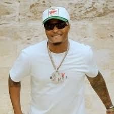 Man in white T-shirt and cap, wearing sunglasses and a gold chain, smiling outdoors against a stone wall