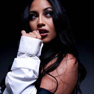 Portrait of a woman with long dark hair in a white top, looking over her shoulder against a black background