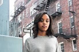 Woman standing on a city sidewalk in front of brick buildings and fire escapes, wearing a gray sweater.