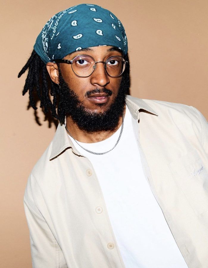 Portrait of a man wearing a blue bandana, glasses, and a cream shirt against a tan background