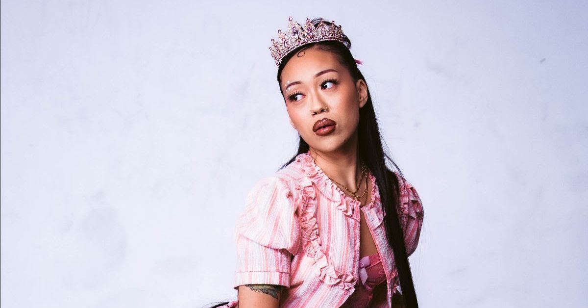 Woman in a pink floral dress and crown poses against a plain white background, looking off to the side