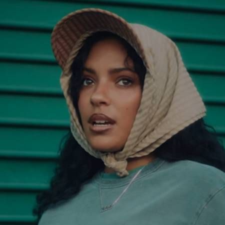 Woman in a beige headscarf and teal sweatshirt, standing before a green corrugated wall.
