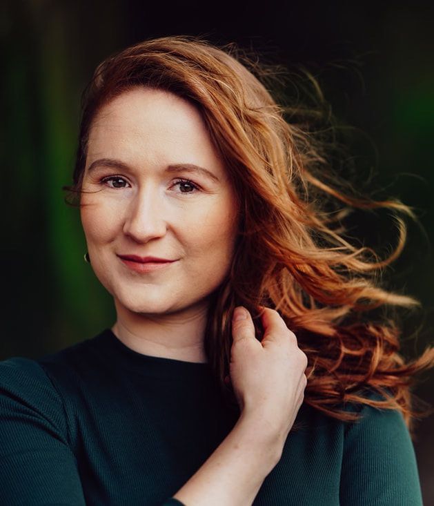 Portrait of a red-haired person in a dark top, smiling softly with hair blown over one shoulder.
