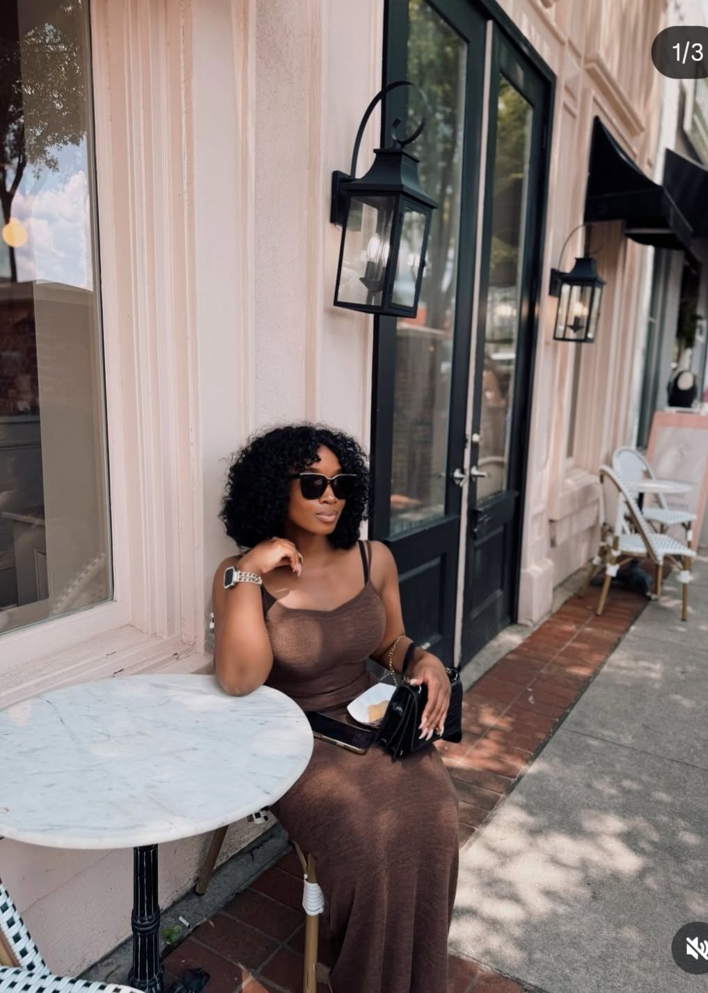 Woman in a brown dress sitting at a café table outside a storefront, wearing sunglasses.