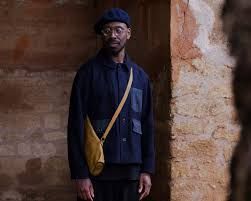 Man standing against a textured wall, wearing a dark jacket, blue beret, and yellow crossbody bag.