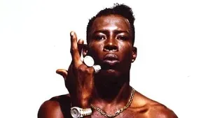 Portrait of a shirtless man against a white background, holding a hand with rings near his face.
