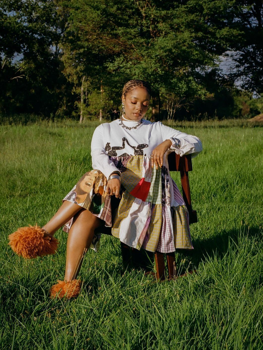 Person seated in a grassy field wearing a white top and patterned skirt, with orange fluffy shoes, in sunlight
