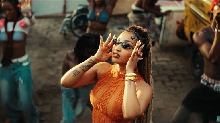 Woman in an orange dress adjusting sunglasses while dancing outdoors amid a lively street crowd.