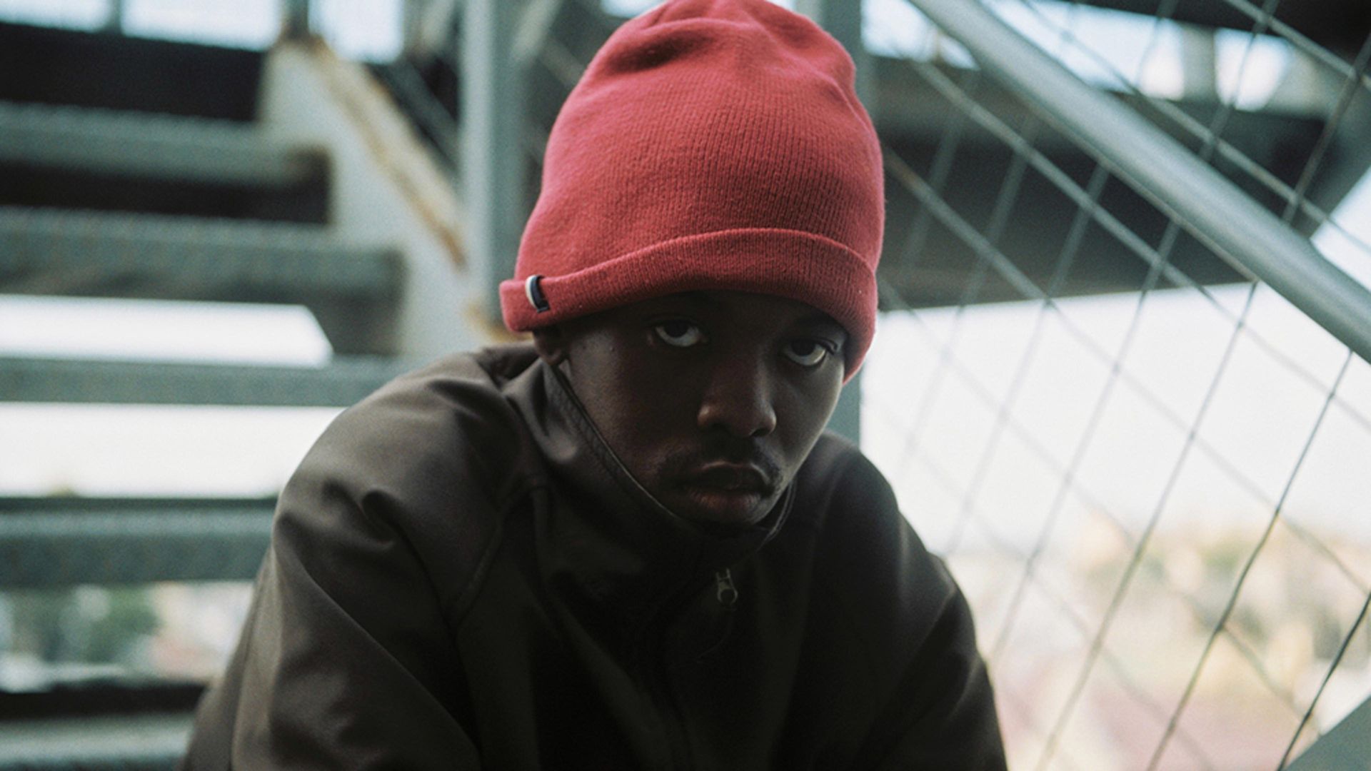 Person in a red beanie and dark coat sitting by a windowed industrial structure, looking at the camera