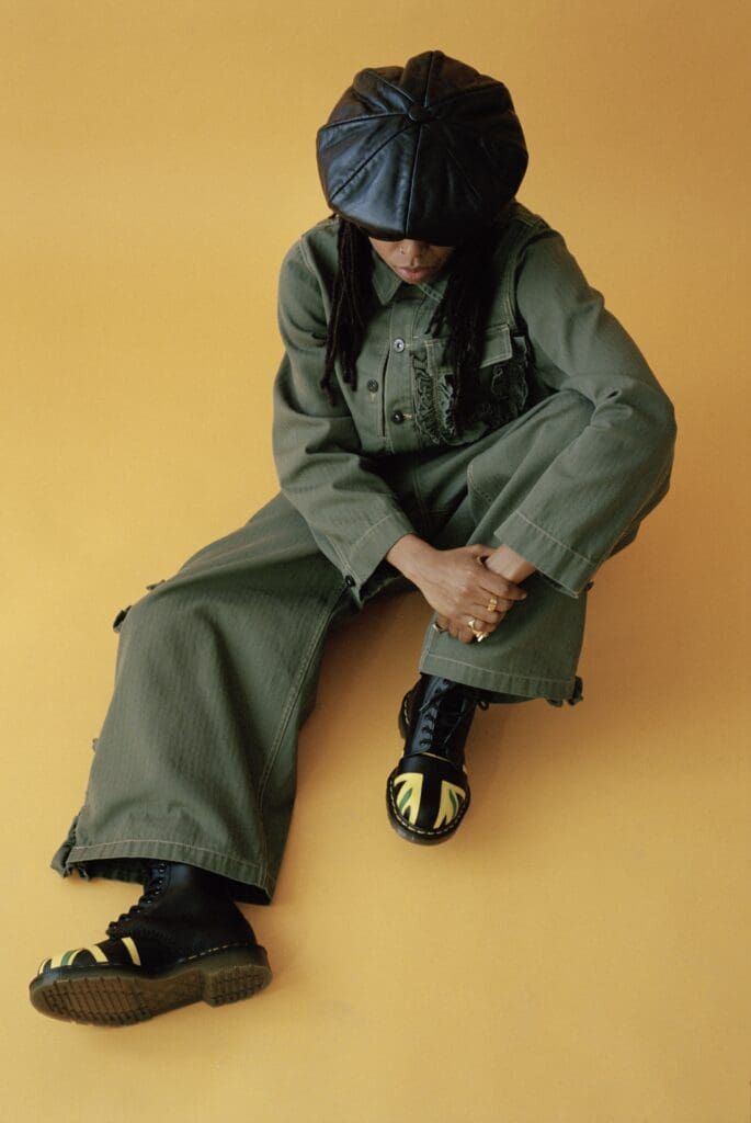 Person in green clothing and black head covering sitting on a yellow background, looking down