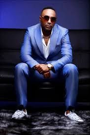 Man in a light blue suit and sunglasses seated on a black couch against a dark blue wall