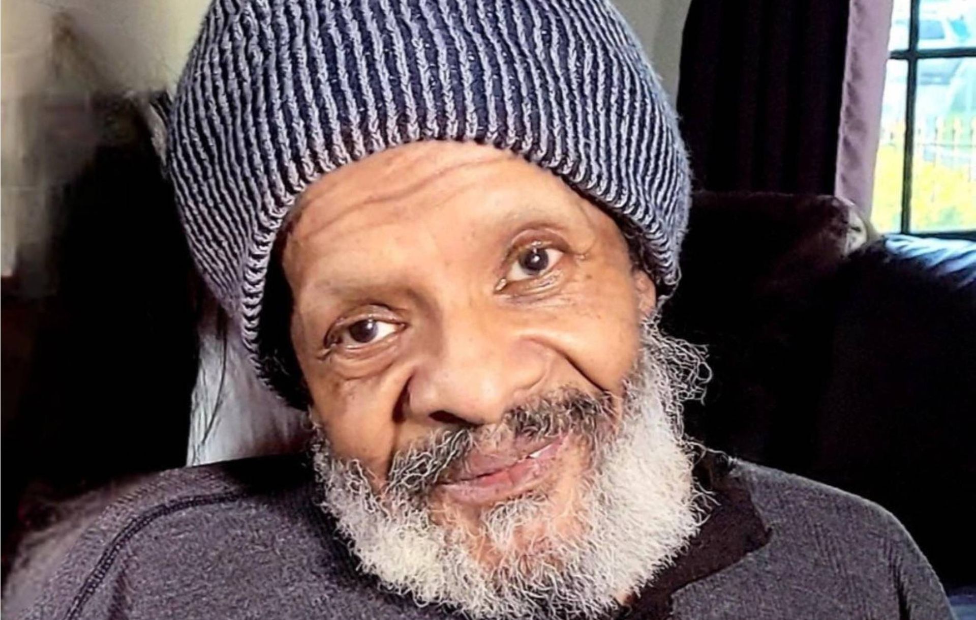 Smiling man in a knit beanie, seated indoors near a window.