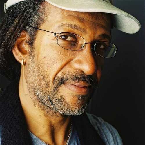 Close-up portrait of a man wearing glasses and a light hat, with a thoughtful expression against a dark background