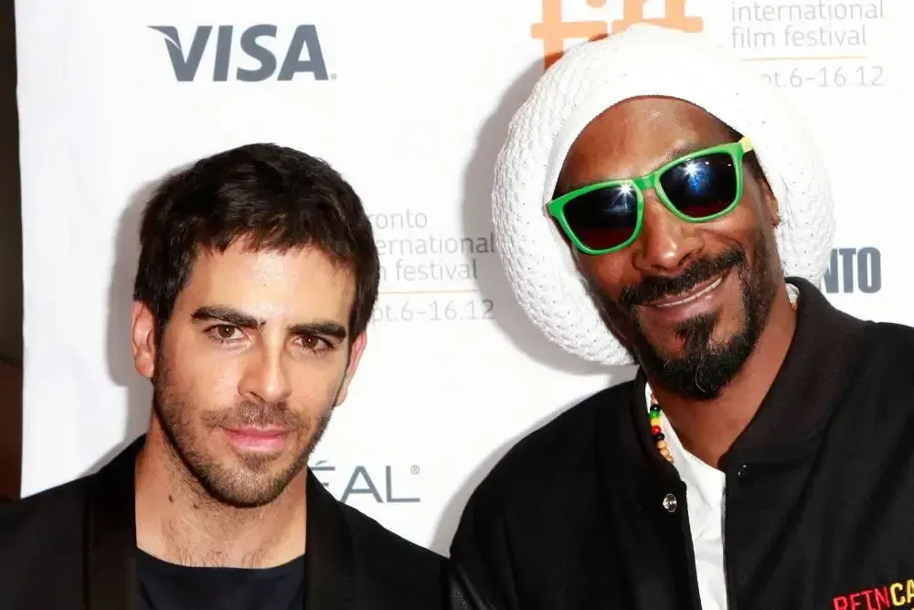 Two men posing at a film event, one wearing green sunglasses and a white headwrap, in front of sponsor logos