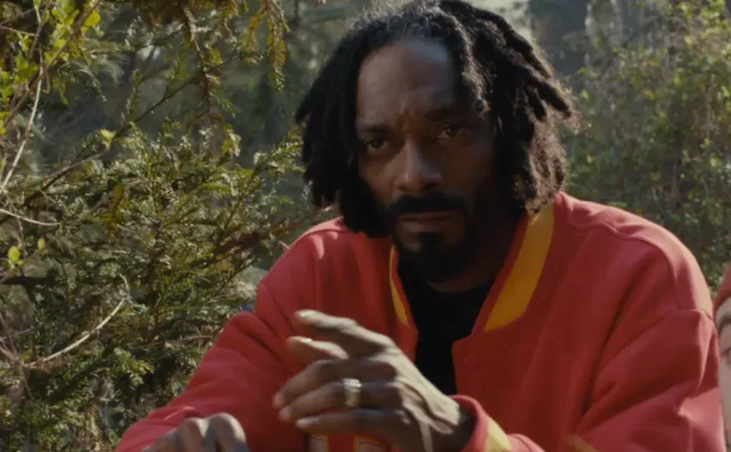 Man in a red robe sitting outdoors among trees, leaning forward with hands clasped.