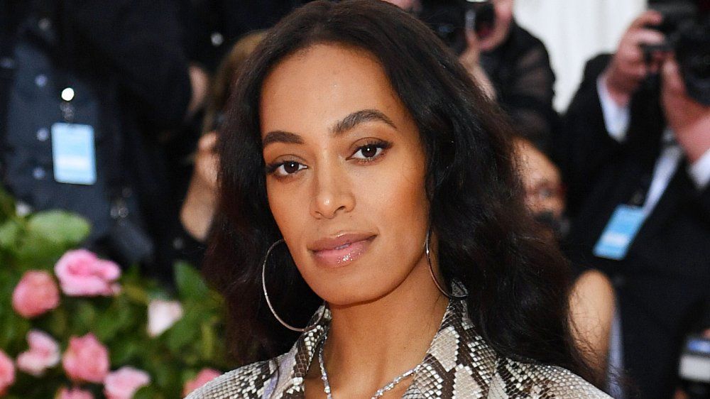 Woman posing at a red carpet event, wearing a patterned outfit and hoop earrings, with flowers in the background