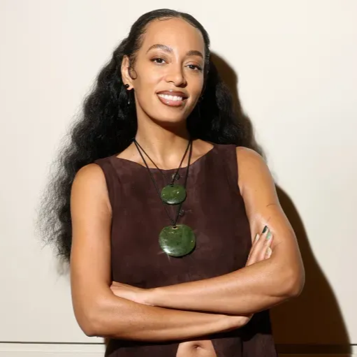 Person in a sleeveless brown top with crossed arms, wearing a green pendant necklace, smiling against a light background.