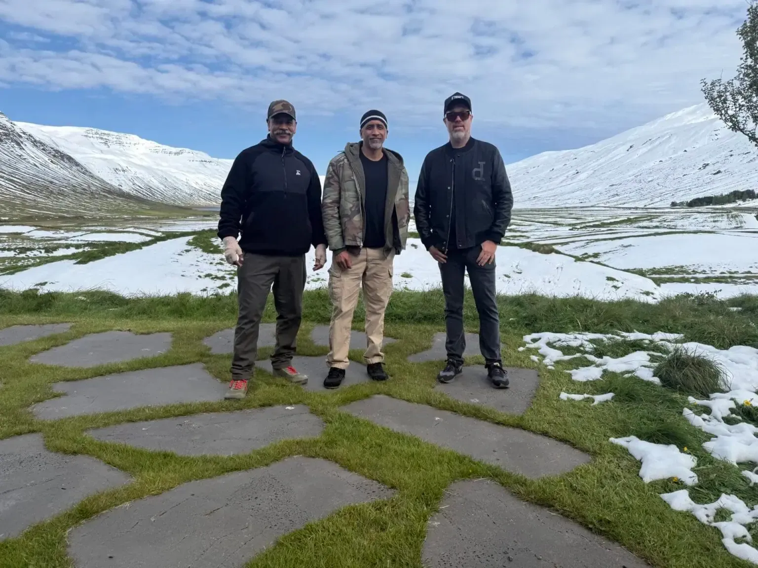 Three people standing on a snowy mountain trail beside a lake under a cloudy sky.