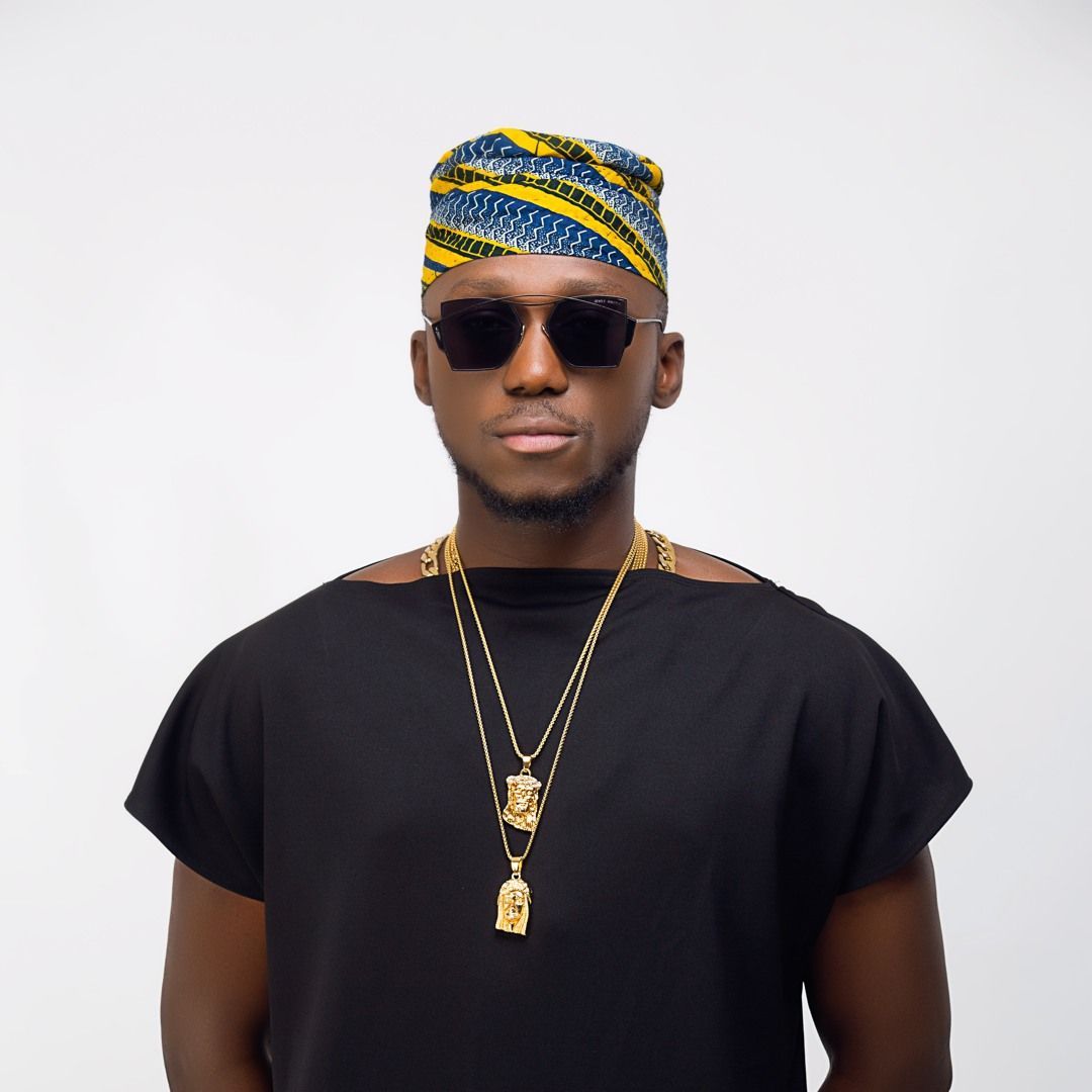 Portrait of a man wearing sunglasses, a patterned headwrap, black shirt, and gold necklaces on a white background