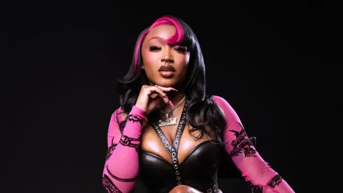Portrait of a person with pink hair, black outfit, and finger on chin against a black background