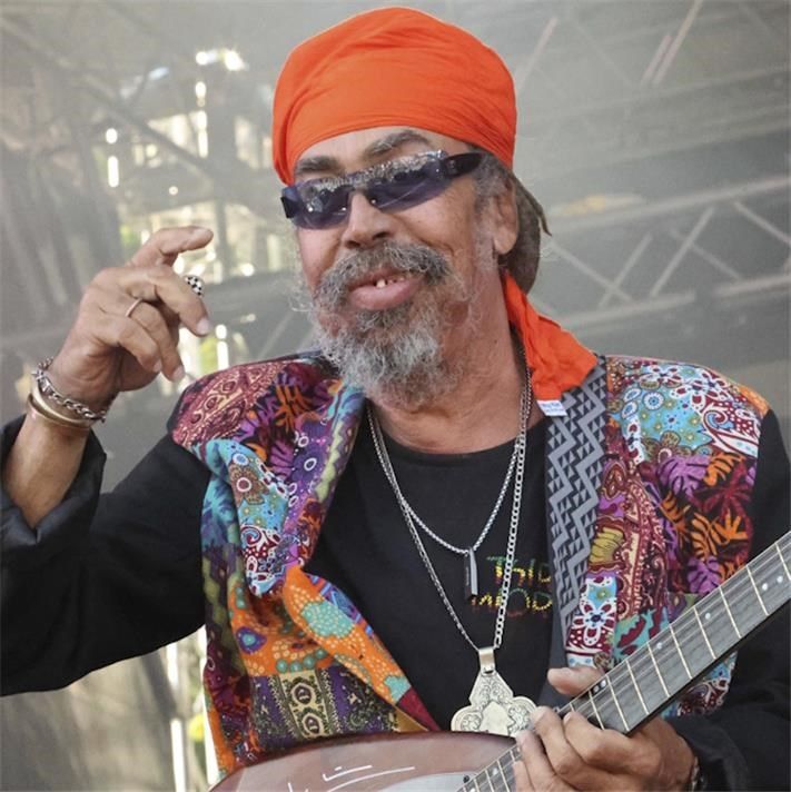 Musician in orange turban and sunglasses performing on stage with a guitar, wearing a colorful vest.