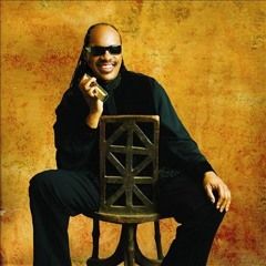 Man seated on a chair, wearing dark clothes and sunglasses, smiling against a warm textured background