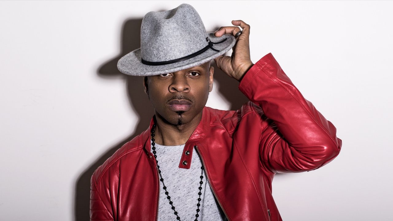 Man in a gray hat and red jacket adjusting the brim against a white background