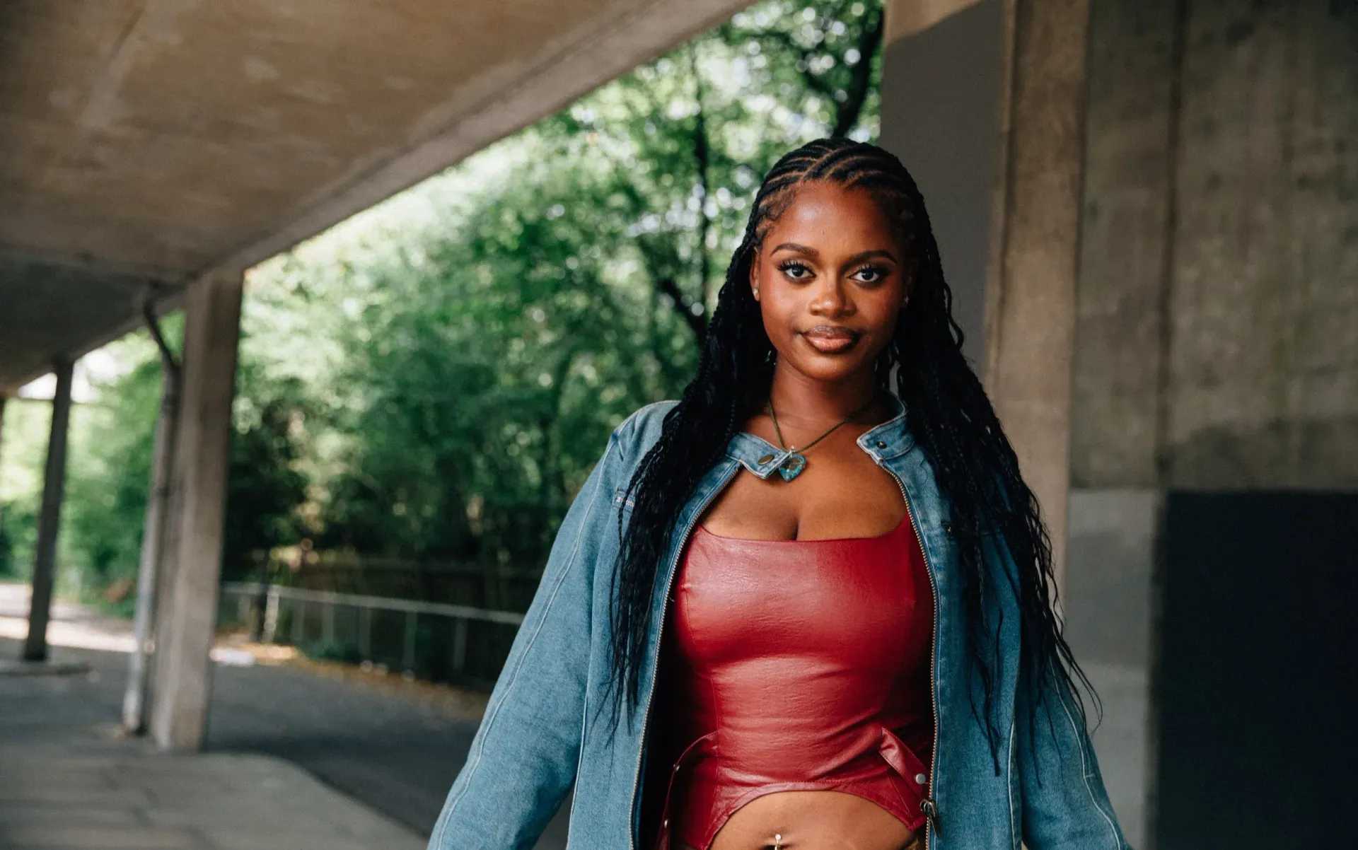 Woman in a red dress and denim jacket standing under a concrete overpass with trees in the background