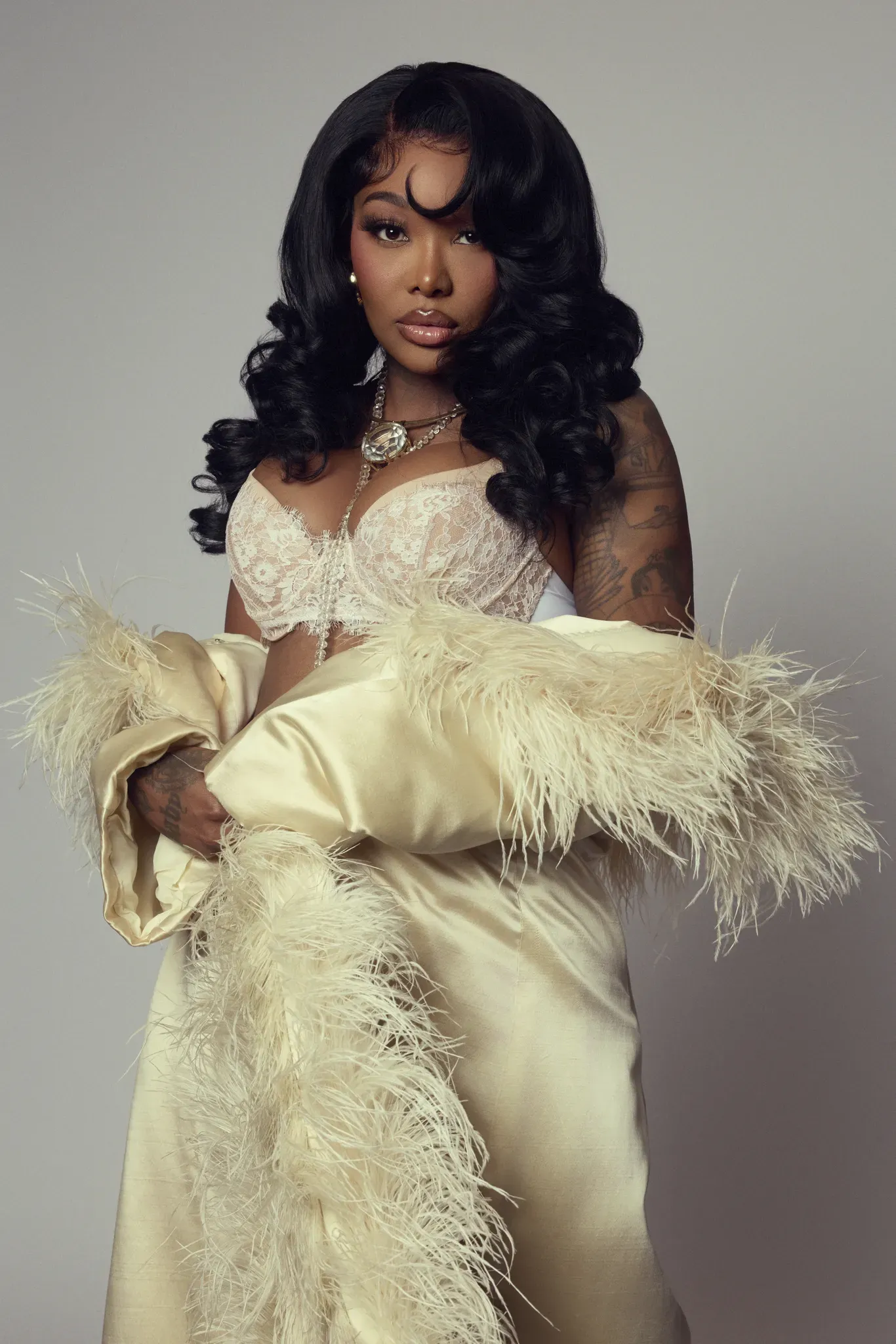 Woman in a cream lace gown and feather boa posing against a gray backdrop.