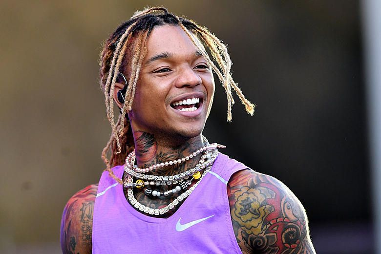 Smiling athlete in a purple Nike tank top with layered necklaces and tattooed arms, standing outdoors.