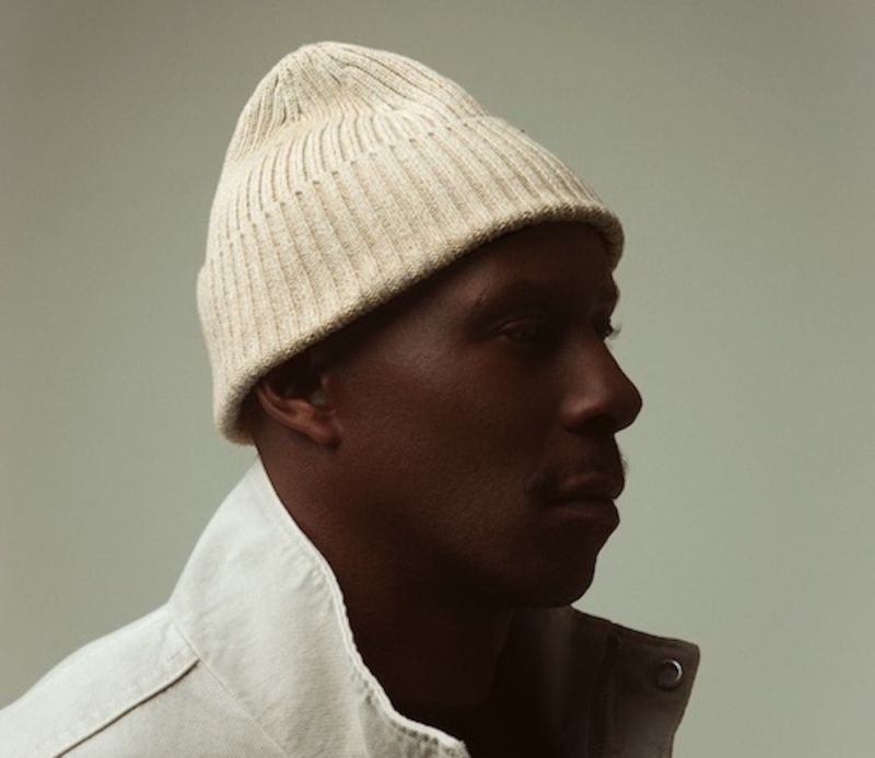 Profile of a person in a cream knit beanie and white jacket against a plain gray background