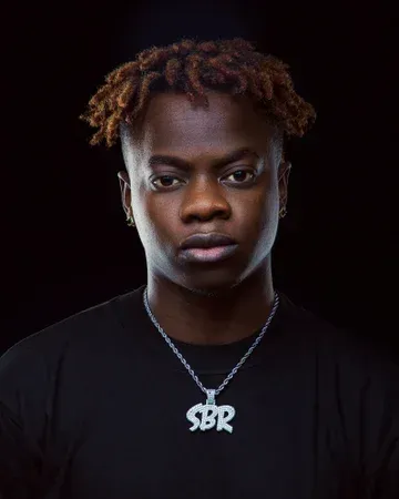 Studio portrait of a person in a black shirt and silver necklace on a dark background, facing forward with a serious expression