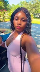 Person taking a selfie outdoors on a sunny street, wearing a light pink top.