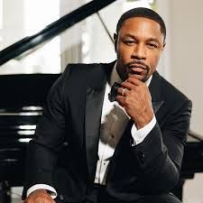 Man in a black tuxedo posing beside a grand piano, resting his chin on his hand