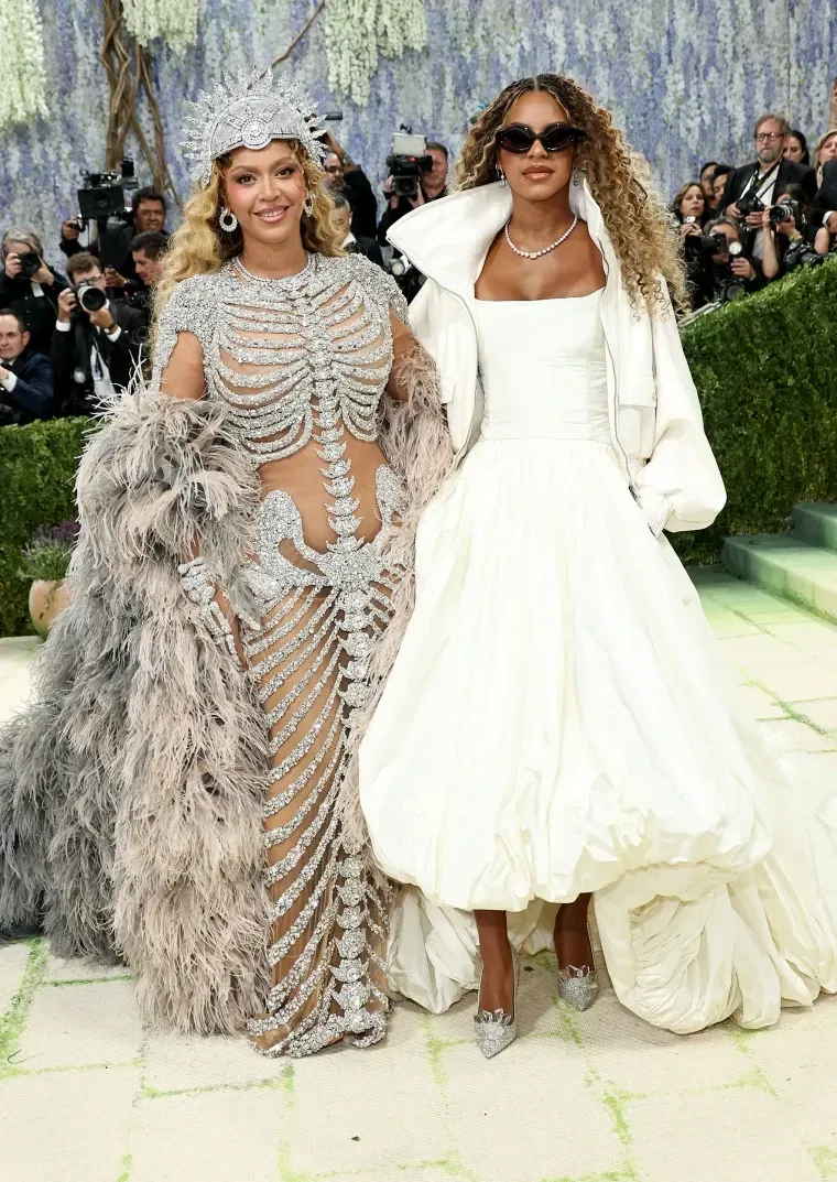 Two women in ornate silver and white gowns posing on a red carpet