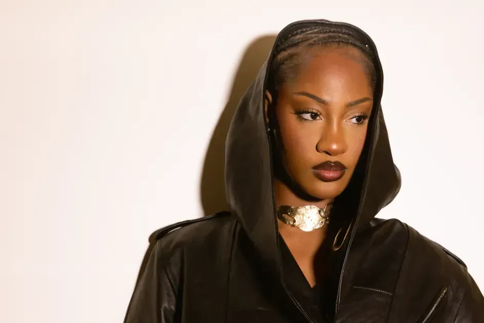 Person in a black hooded jacket with a gold choker, looking to the side against a light background