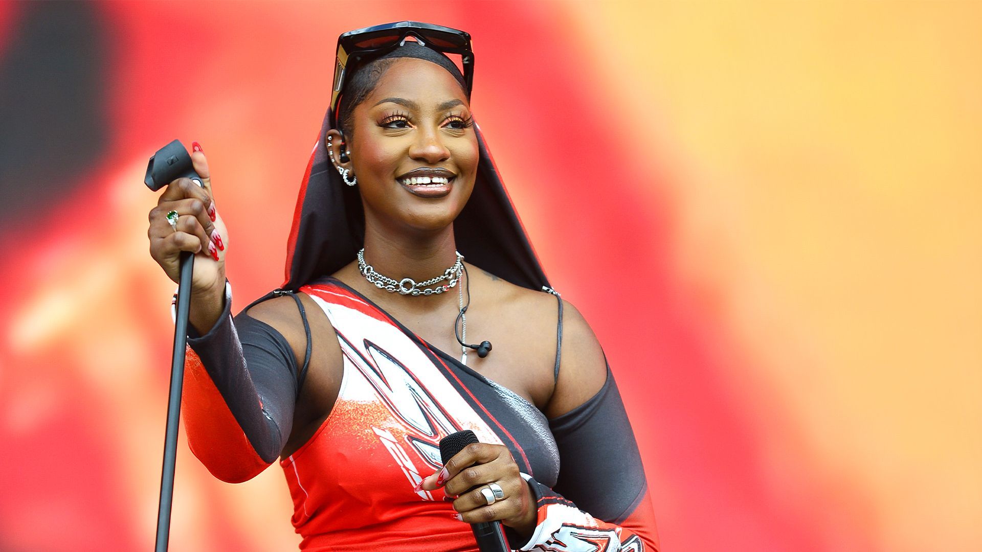 Singer performing onstage, smiling and holding a microphone against a red-orange backdrop.