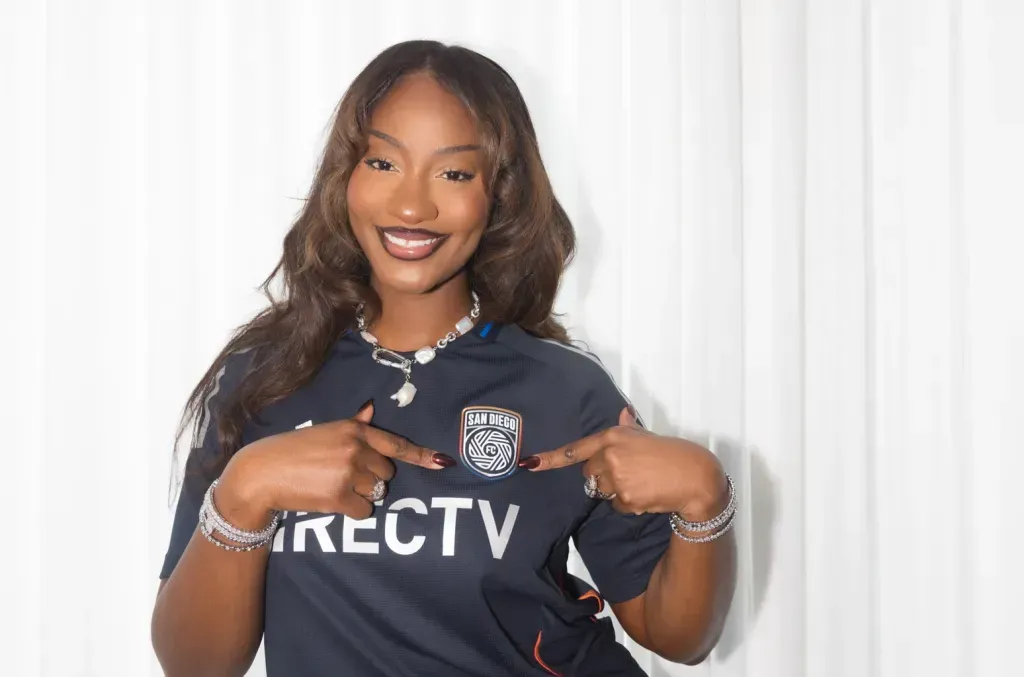 Woman smiling in navy DIRECTV soccer jersey, pointing at the team crest against a white backdrop