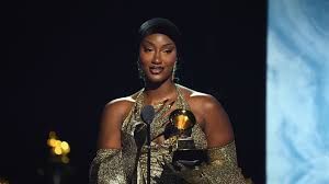 Woman holding a trophy on stage, wearing a gold dress, with microphones and blue-lit background.