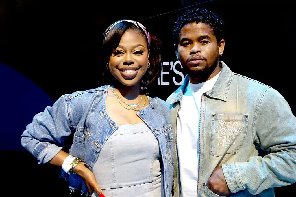 Two people smiling onstage, both in denim jackets, posing side by side against a dark background.