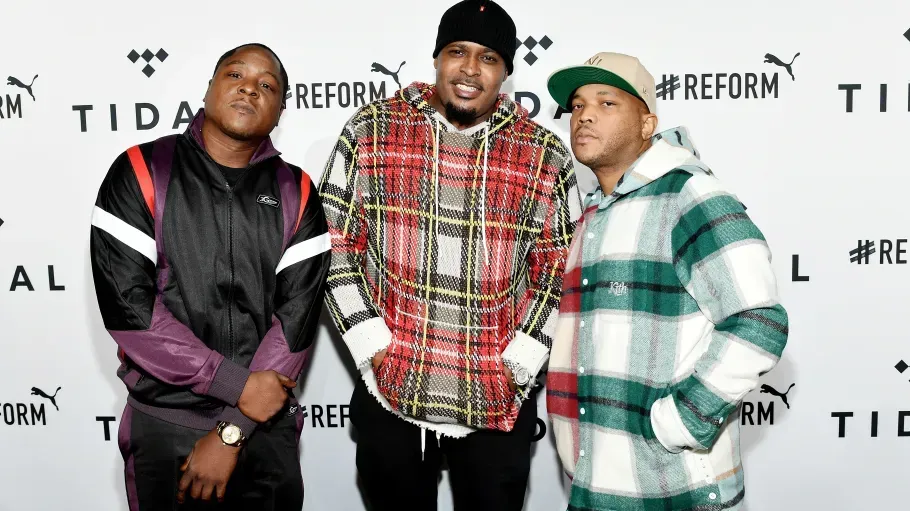 Three men posing at a red-carpet event, wearing black, red plaid, and green plaid outfits.