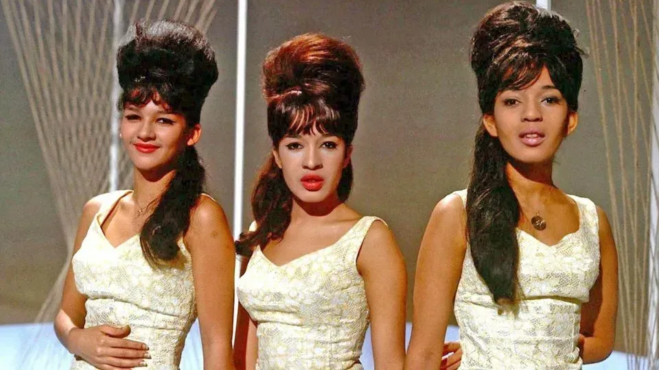 Three women in matching white dresses sing onstage against a warm studio backdrop.