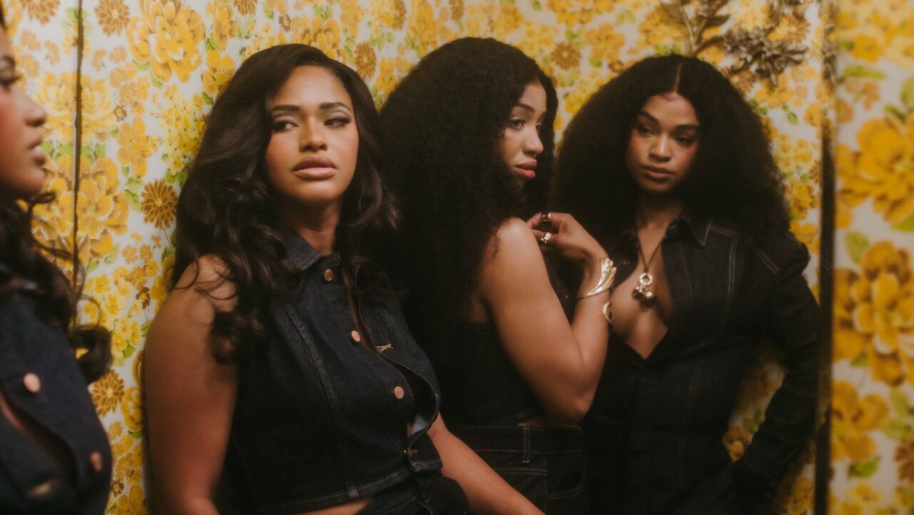 Four people posing against a yellow floral wallpaper backdrop, wearing dark clothing.