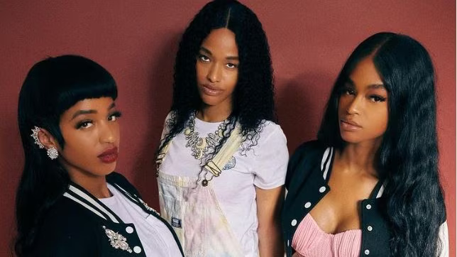 Three people posing against a red background, wearing black and white outfits and jewelry.