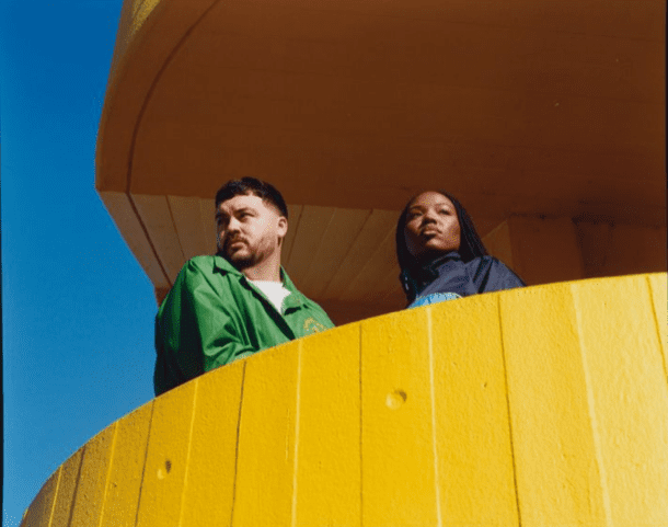 Two people stand on a bright yellow balcony against a blue sky, looking outward.