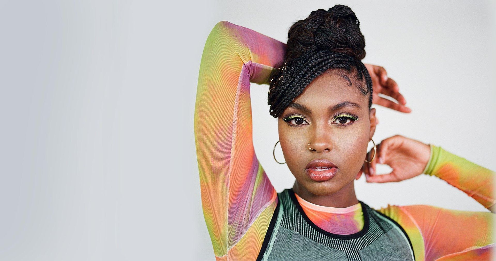 Portrait of a person posing with hand behind head, wearing a colorful mesh top against a light background