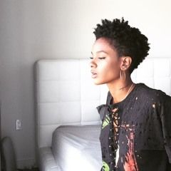 Woman in profile beside a bed, with short curly hair and a black patterned jacket.