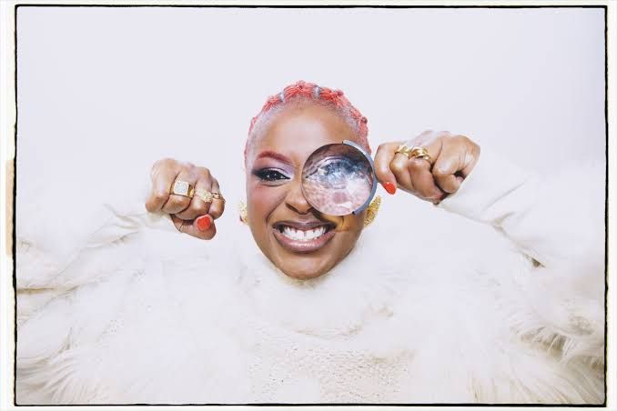 Smiling person in white fluff holds magnifying glasses up to both eyes, with pink hair against a white background