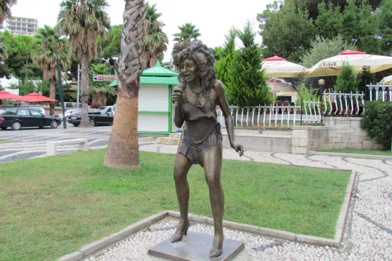 Bronze statue of a standing woman in a park near palm trees and a paved walkway